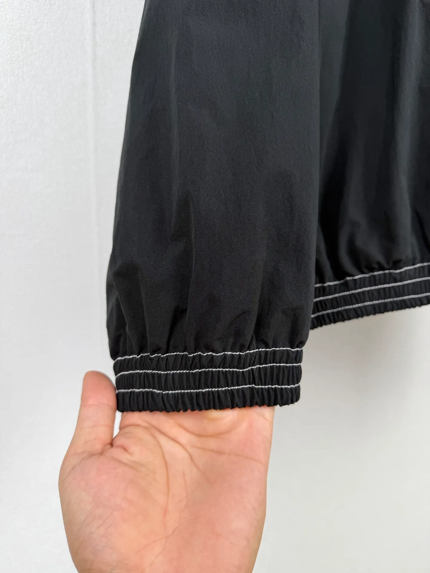 ACNE Studios back letter topstitch hooded jacket is made of fabric processing, contrasting topstitching, printing alignment and combination of elastic components. The fabric is treated with a physical wrinkle process, which does not rely on chemical coatings. It uses mechanical wrinkles to create a disordered texture effect on the surface of the fabric. While the fabric has a relaxed look, it still retains the structural tension of the woven fabric. During the cutting stage, the deformation caused by the folds of the fabric needs to be fully considered to ensure a stable silhouette after the pieces are spliced. The entire product uses contrasting topstitching as the main structural decoration. The seams of the cap, shoulder seams, cuffs, and hem are all exposed with contrasting topstitching. There are arc turns in different parts of the cut pieces. When sewing, it is necessary to control the fabric feeding rhythm and maintain a uniform stitch density. The elastic area of ​​the hem is sewn with multiple layers of materials. The layers of fabric, elastic bands, and hemming pieces are overlapped. It is easy for the line to shift and the fabric to arch, which requires high precision in the lathe operation. The Retro character printing on the back is the visual core of the entire garment. Multiple groups of characters are arranged in layers, giving it a worn and broken texture. The printing must ensure that the layer is strong and washable, but cannot cover up the details of the incomplete retro characters. At the same time, the printing positioning needs to be aligned with the middle seam of the back of the garment to avoid tilting and offset of the printing. The cuffs and hem are both elastic pleated structures. When sewing, you need to control the density of the fabric pleats to ensure a uniform and natural arc after shrinkage. The hood is spliced ​​into pieces and relies on multiple open threads to separate and shape it, ensuring that the hat is natural and three-dimensional and will not collapse against the face when worn. After all the sewing processes are completed, the garment needs to be ironed and finished to unify the wrinkles of the entire fabric and remove excess creases caused by the production process, finally presenting the complete Retro relaxed garment effect. Size: 46 48 50 52